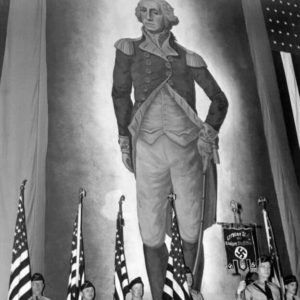 A color guard holding American flags and a banner inscribed with the Nazi swastika stands before an immense portrait of George Washington at the German American Bund's rally at Madison Square Garden.